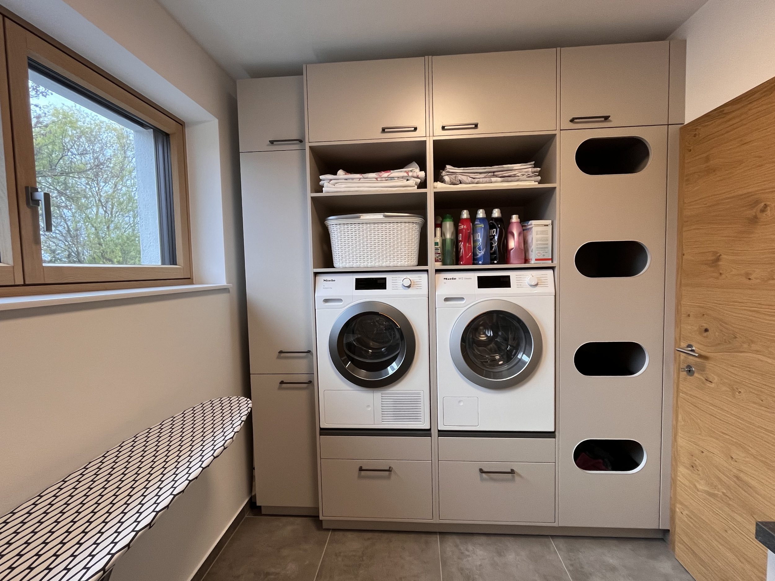 Waschküche mit erhöhter Waschmaschine und Wäscheeinwurf, grau mit schwarzen Griffen